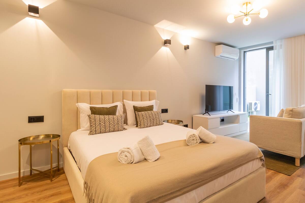 A modern hotel room with a large bed, a TV, a sofa, and a dining area.