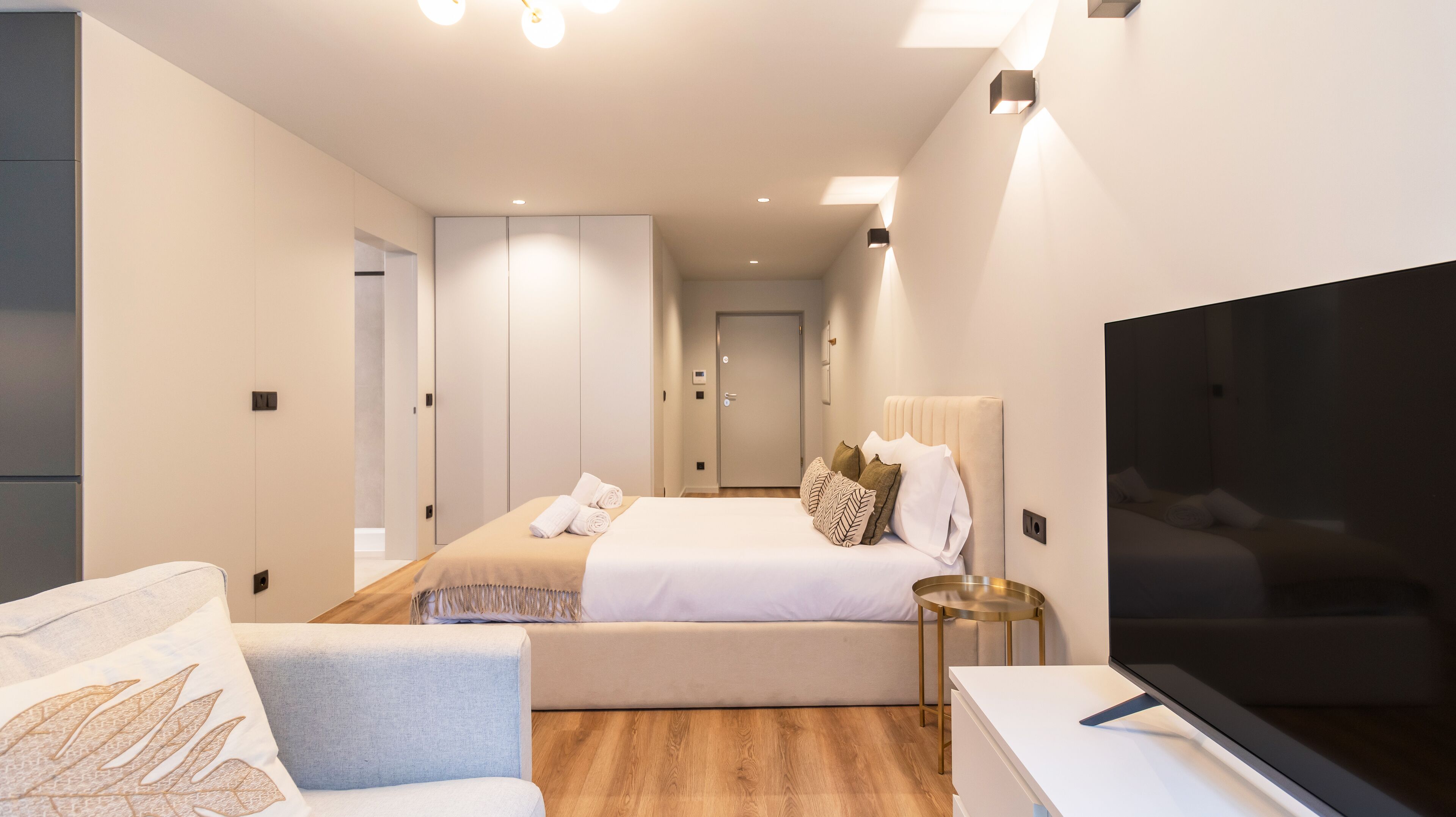 A modern hotel room with a bed, a TV, a sofa, and a bedside table.