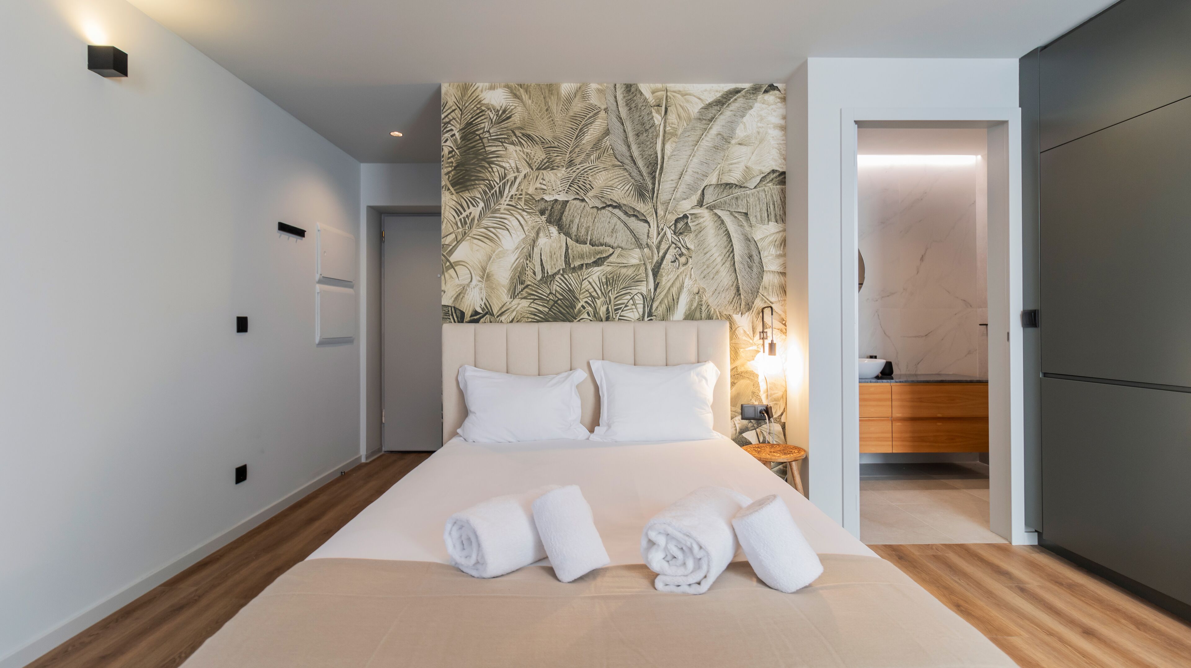 Een moderne hotelkamer met een groot bed, een hoofdeinde met een tropisch bladmotief, een nachtkastje met een lamp en uitzicht op de badkamer.