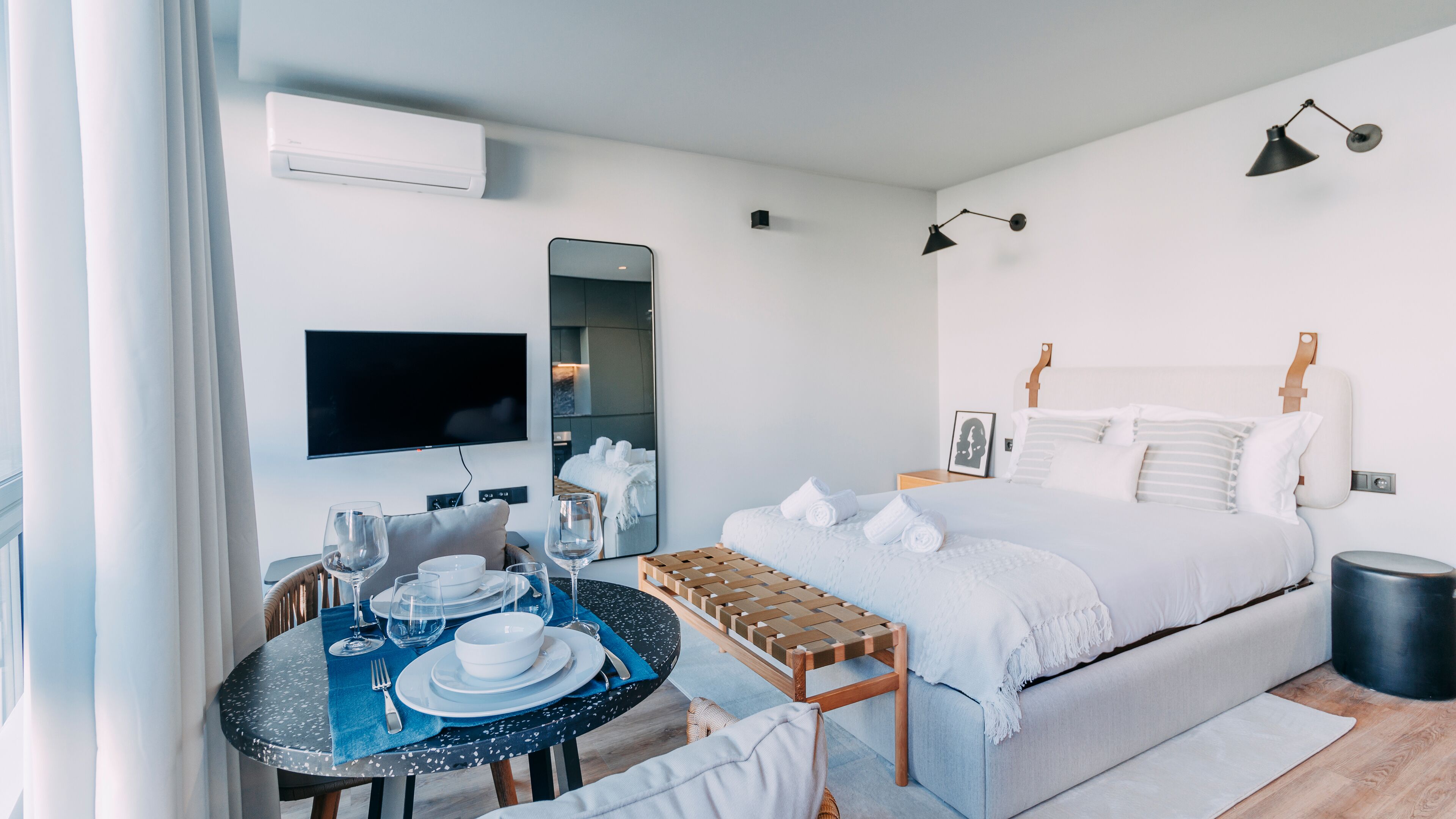 A modern hotel room with a bed, a TV, a dining table set for two, and a small seating area.