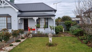 A two-story house with a gabled roof, a front porch with a white lace trim, and a small garden with various plants and a decorative stone.
