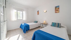A room with two beds, a window with curtains, a small table with a lamp, and two wall-mounted paintings.