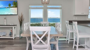 Una cocina moderna con armarios blancos, una mesa de comedor de madera y sillas. Hay un ventanal con vista al océano.