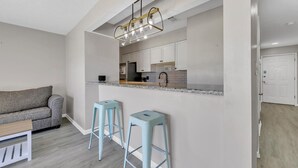 Una cocina moderna con armarios blancos, encimera de mármol y una lámpara colgante. Hay dos taburetes azul claro en la barra y un sofá gris en la sala de estar.
