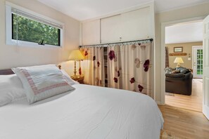 Ein Schlafzimmer mit Bett, Nachttisch, Lampe und Fenster mit Blick auf Bäume.