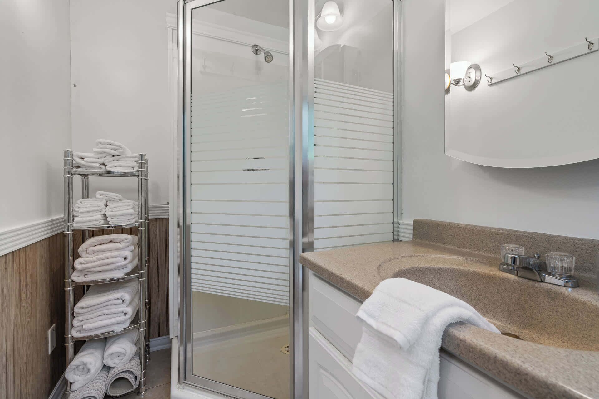 A bathroom with a glass shower enclosure, a towel rack with folded towels, a sink with a mirror, and a towel on the counter.