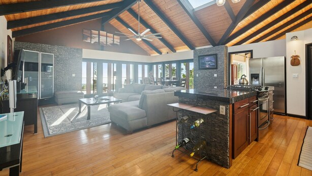 A spacious living area with a high ceiling, a stone fireplace, and a modern kitchen with stainless steel appliances. - Pu'ukala Sunset Estate W/ Amazing Ocean Views! 3 Bedroom Home by RedAwning (Kailua-Kona)