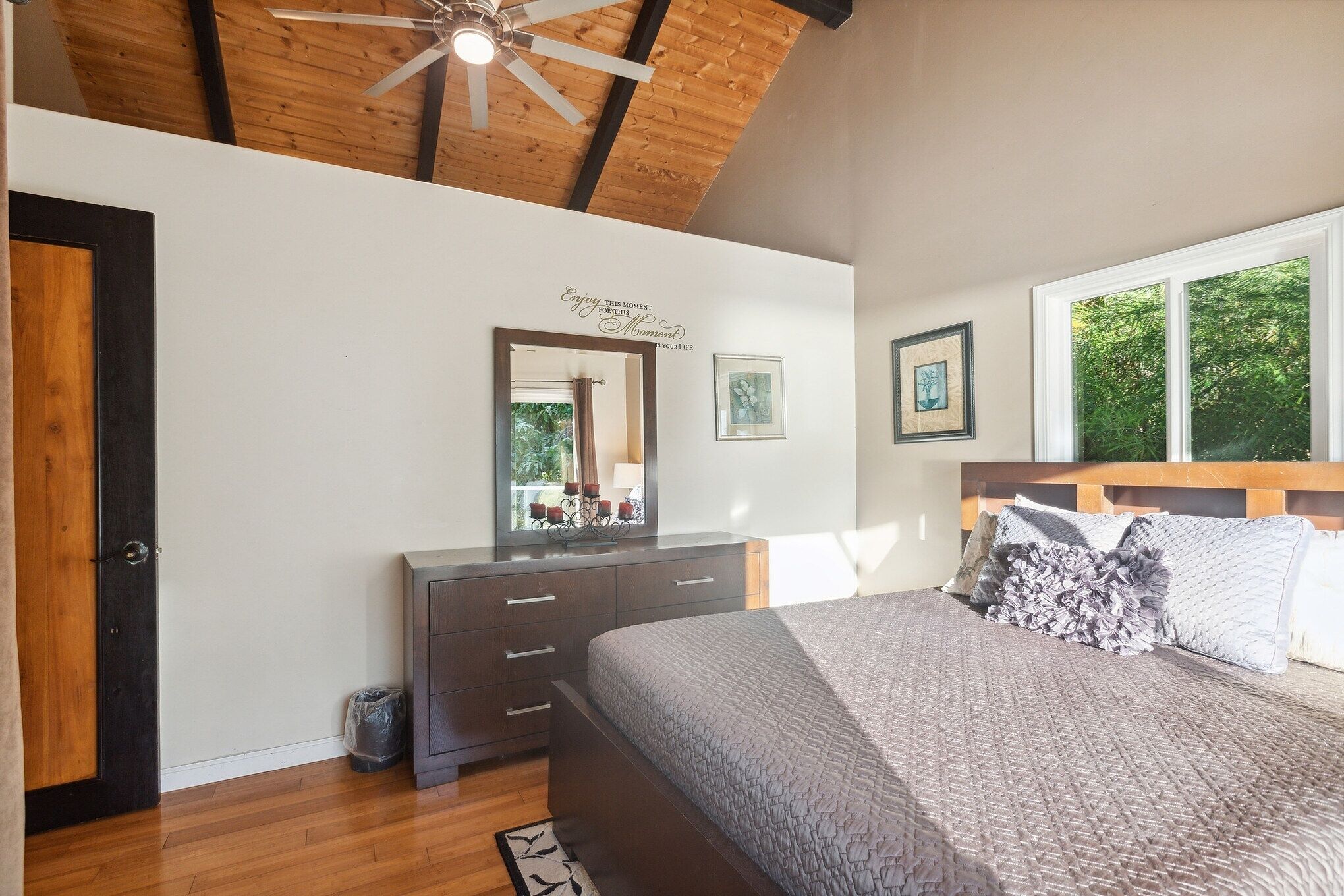 A bedroom with a wooden ceiling, a bed with a patterned bedspread, a dresser with drawers, a mirror, and framed artwork on the wall.