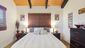 A bedroom with a wooden ceiling, a large bed with white bedding, two bedside tables with lamps, a red vase, and artwork on the walls. - Charming Pu'ukala Sunset - Near Hiking And Golf 1 Bedroom Home by RedAwning (Kailua-Kona)