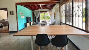 Una larga mesa de madera con dos sillas negras, un televisor sobre un mueble y vistas a un patio exterior a través de una ventana con barrotes.
