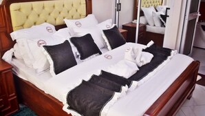 A neatly made bed with white and black bedding, a wooden headboard, and a nightstand with a lamp. - MATCHEU PALACE HÔTEL  (Yaoundé)
