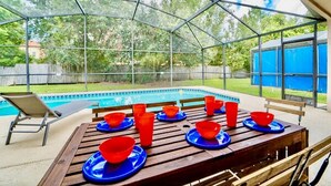Un espacio al aire libre techado con piscina, una mesa de madera con platos azules y cuencos rojos, y una silla.