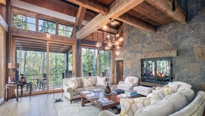 A spacious living room with a stone fireplace, wooden beams, and large windows offering a view of the forest. - Robber's Roost- Sprawling, Ski In/Out, Arcade, Pool Table, & More! (Mountain Village)