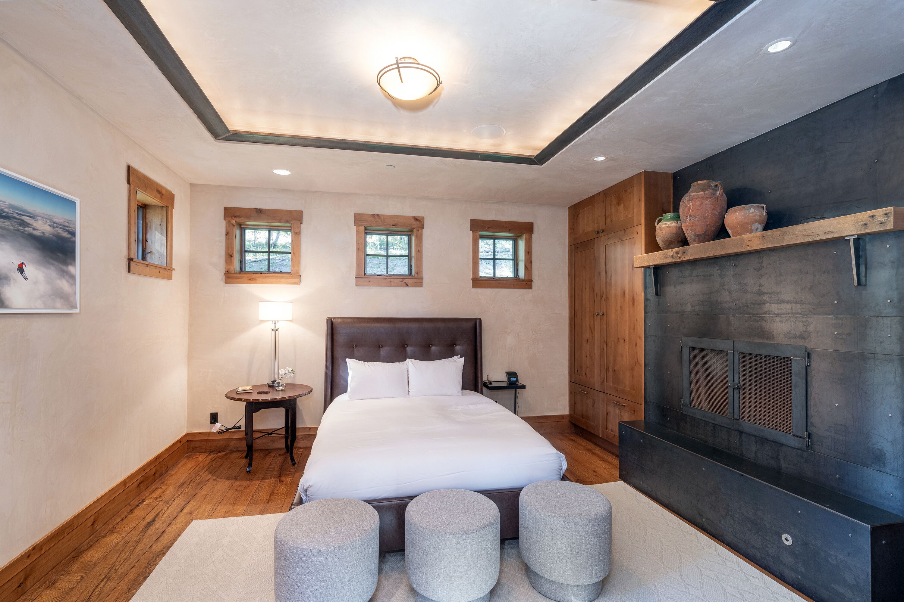A bedroom with a large bed, a bedside table, a lamp, three round ottomans, and a fireplace.