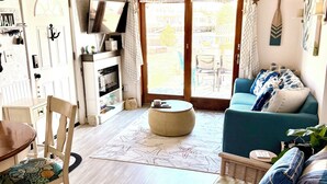 Een gezellige woonkamer met houten vloer, een groene bank, een rieten stoel en een eettafel met stoelen.