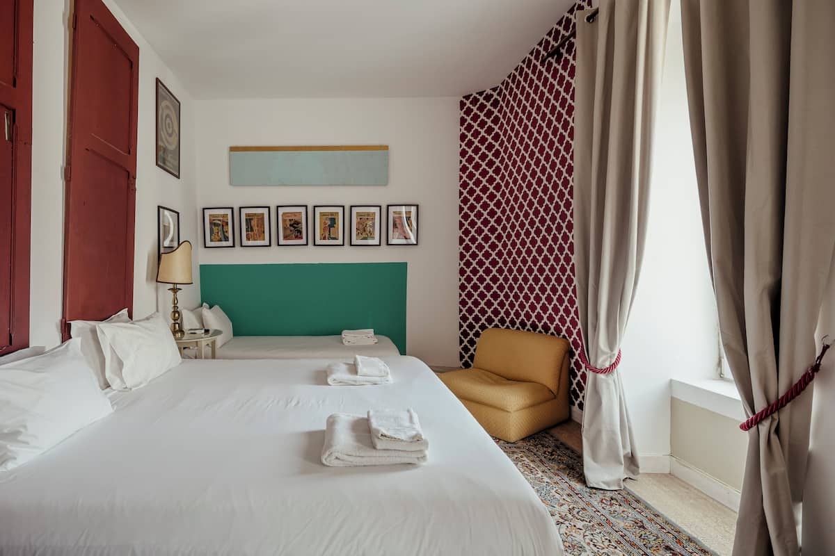 A hotel room with a large bed, a chair, a small table with a lamp, and a window with curtains.