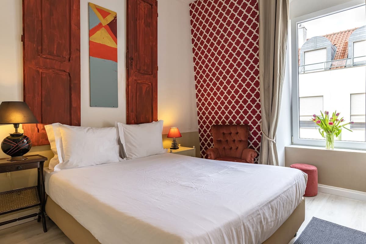 A hotel room with a large bed, a nightstand with a lamp, a chair, a window with a view of buildings, and a wall with a geometric painting.