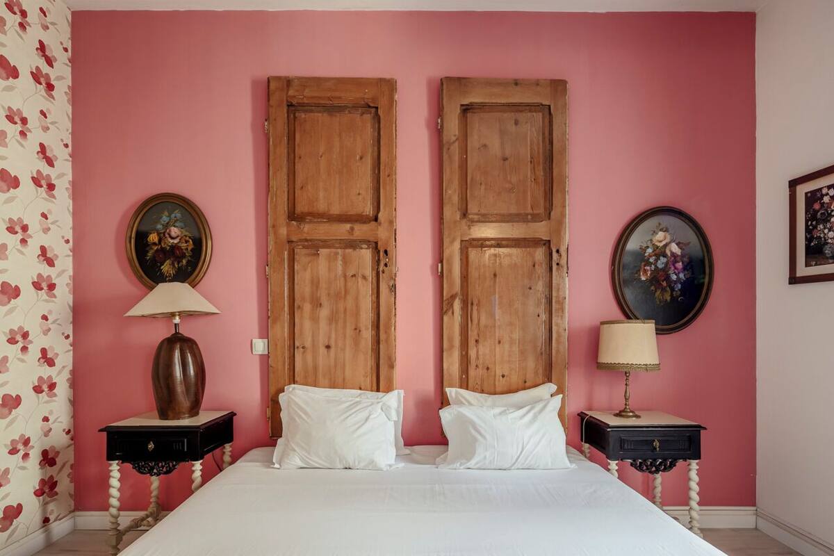 A bedroom with a pink wall, two wooden doors, a bed with white linens, two bedside tables with lamps, and floral wallpaper on the left.