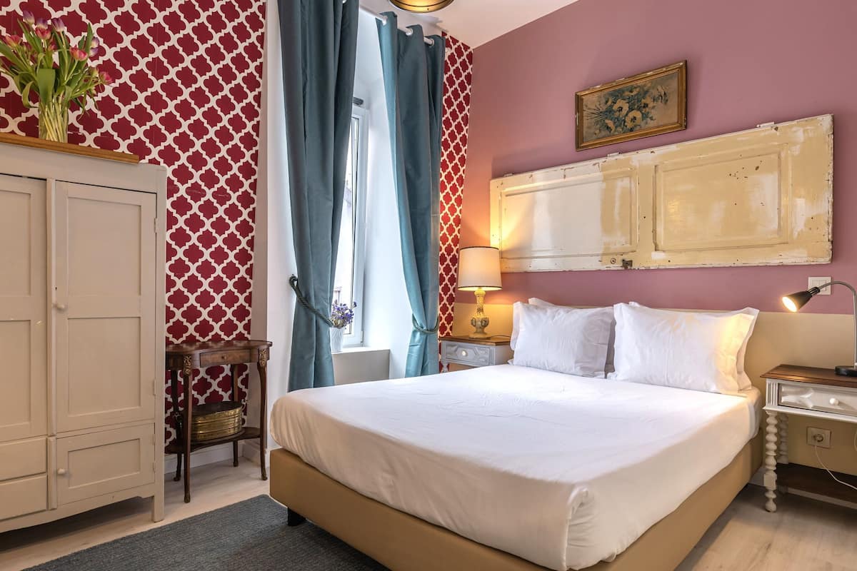 A bedroom with a bed, a wardrobe, a nightstand, a lamp, and a window with curtains.