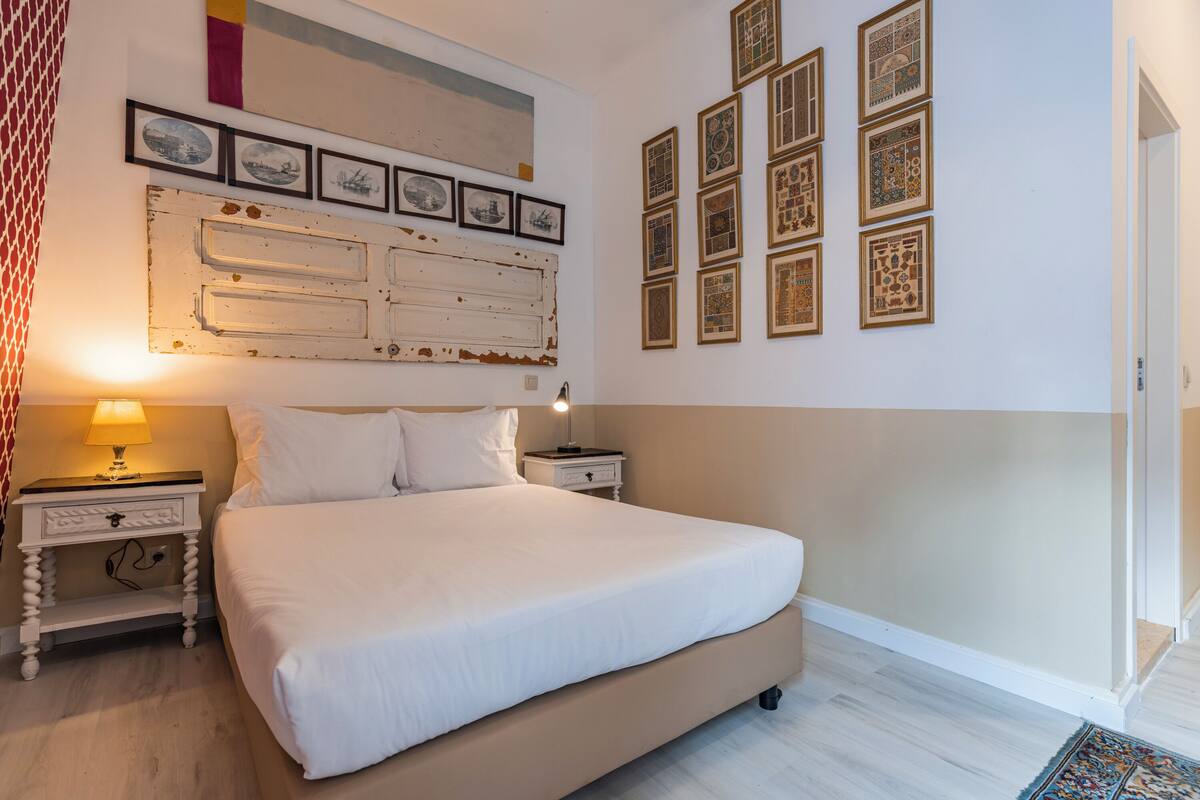 A bedroom with a large bed, two bedside tables with lamps, a wall-mounted headboard, and a series of framed pictures.