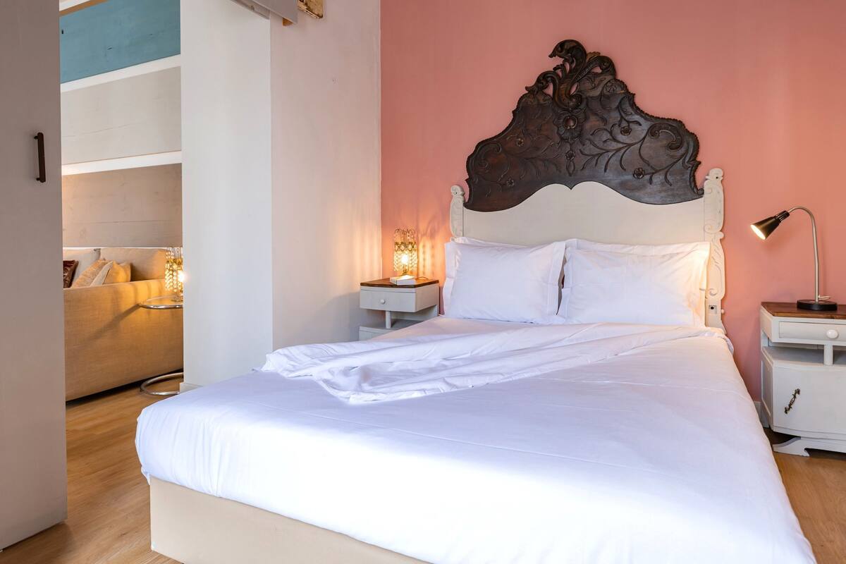A neatly made bed with white linens, a wooden headboard, bedside tables with lamps, and a pink wall.