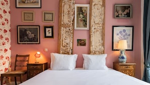 Un lit bien fait, avec du linge de lit blanc, une chaise en bois, une table de chevet avec une lampe, et plusieurs tableaux encadrés accrochés au mur.