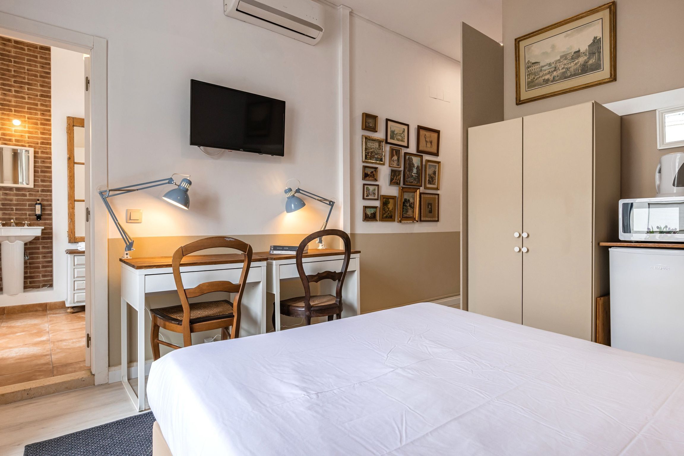 A hotel room with a bed, desk, chairs, a TV, a refrigerator, and a wall with framed pictures.