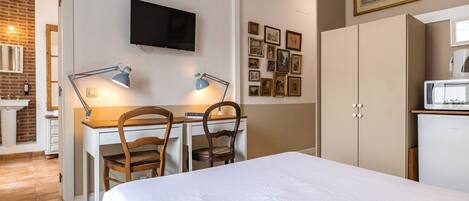 A hotel room with a bed, desk, chairs, a TV, a refrigerator, and a wall with framed pictures.