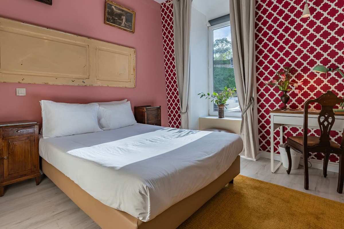 A bedroom with a bed, wooden nightstands, a desk with a chair, a window with curtains, and a patterned wall.