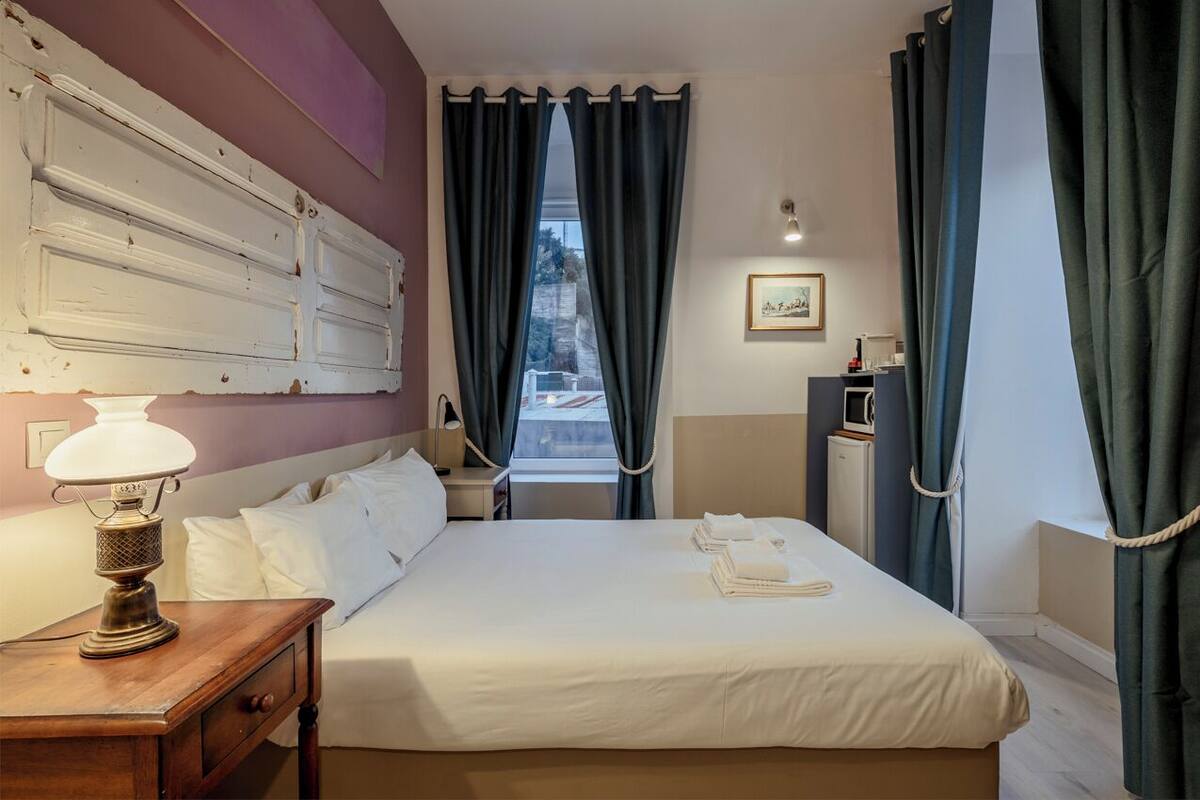 A hotel room with a bed, a bedside table with a lamp, a desk with a chair, a small cabinet, and a window with curtains.