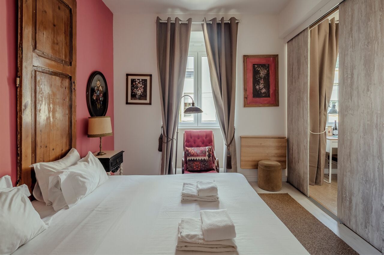 A hotel room with a large bed, a wooden wardrobe, a chair, a small table with a lamp, and a window with curtains.