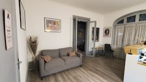 A living room with a grey sofa, a small table, and a chair. There are framed pictures on the walls and a window with curtains.
