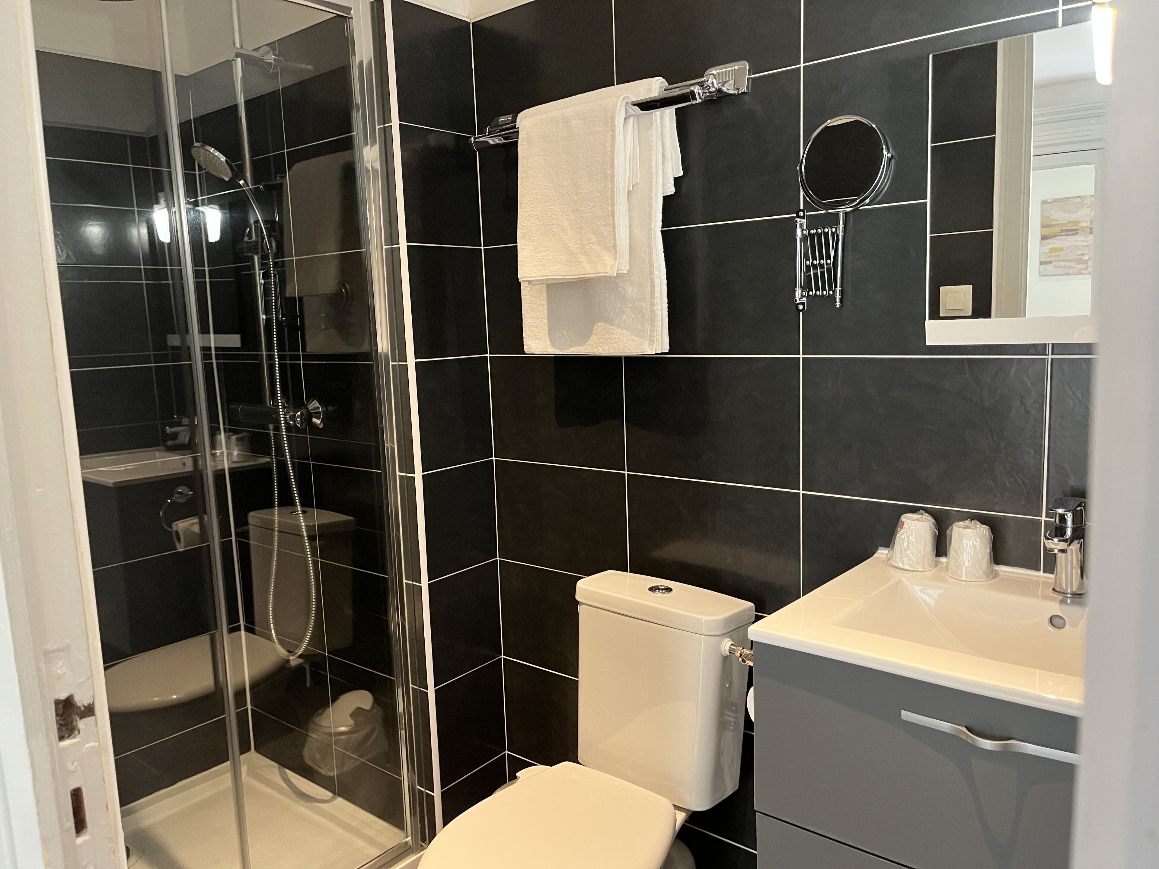 Ein modernes Badezimmer mit schwarzen Fliesen, einer Glaskabine zur Dusche, einer Toilette und einem Waschbecken mit Pflegeprodukten.