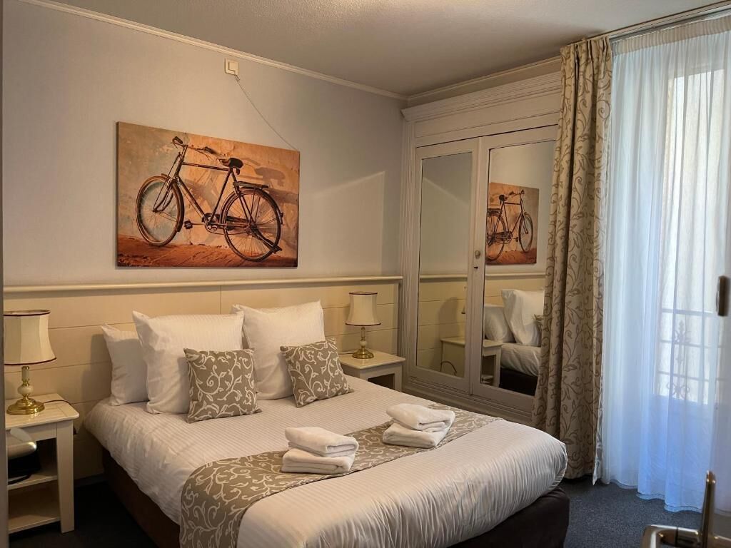 Quarto de hotel com cama, almofadas, mesa de cabeceira com abajur, espelho e pintura de uma bicicleta.