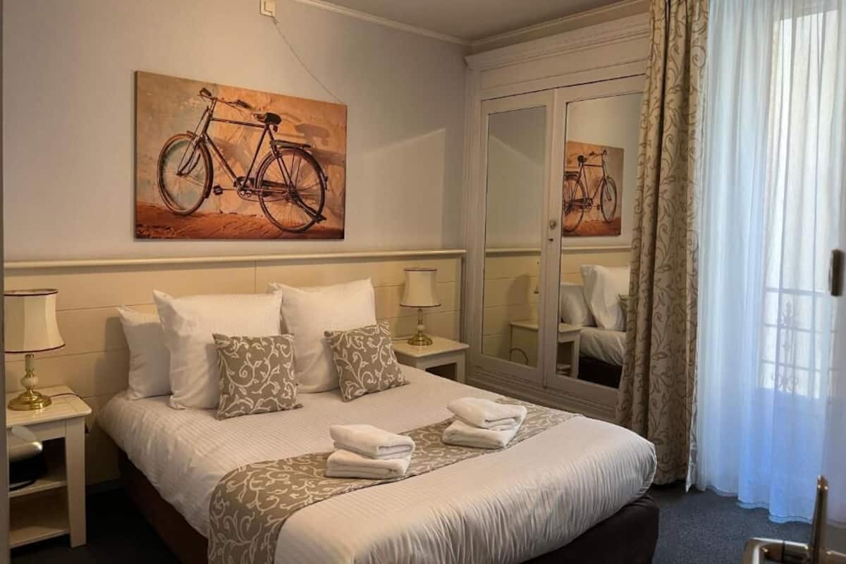 A hotel room with a bed, pillows, a nightstand with a lamp, a mirror, and a painting of a bicycle.