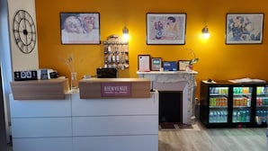 A reception desk with a 'Bienvenue' sign, framed artwork, a clock, and a minibar.