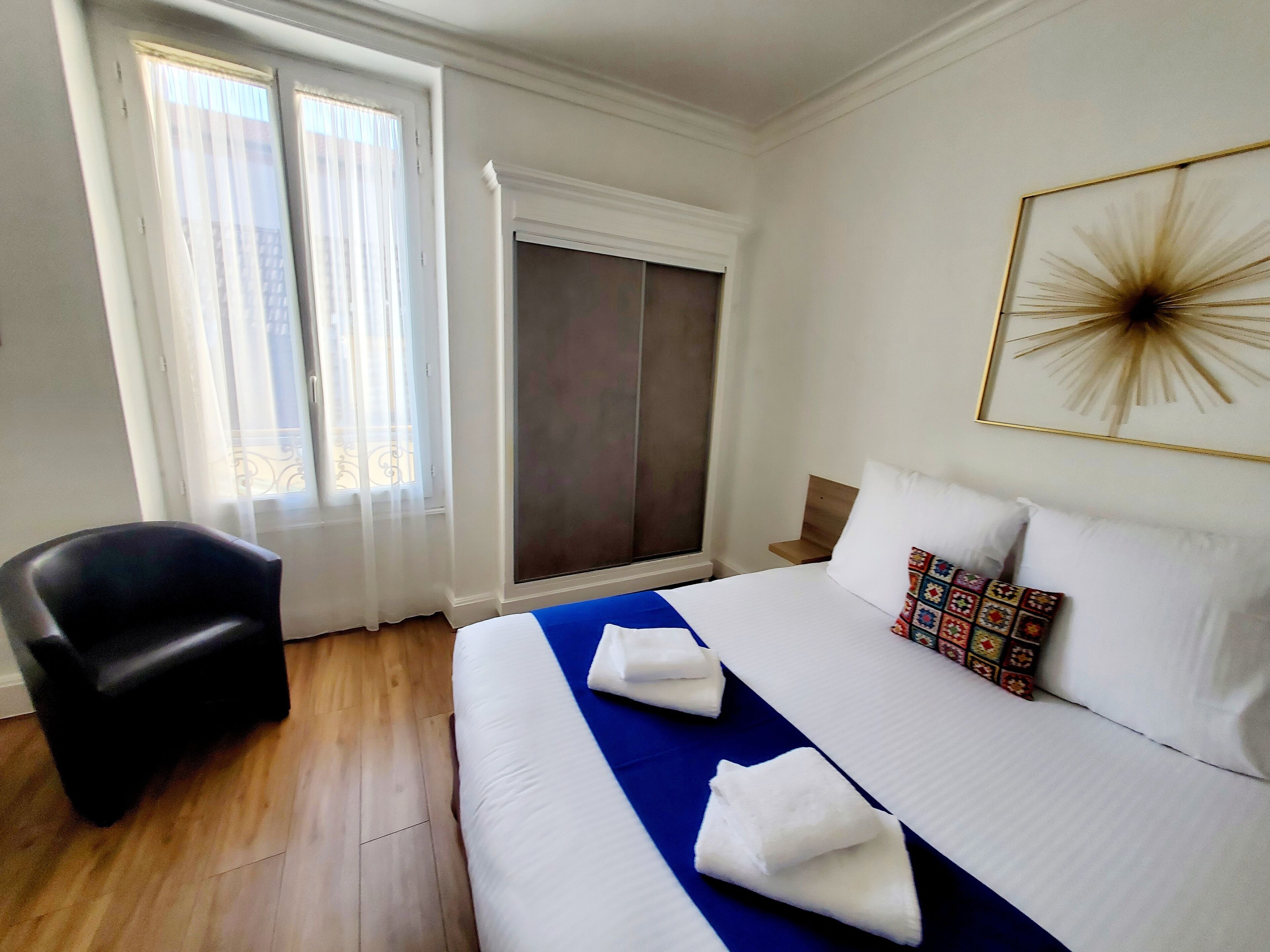 Ein Hotelzimmer mit Bett, Sessel, Fenster mit Vorhängen, Tür und Wandbild.