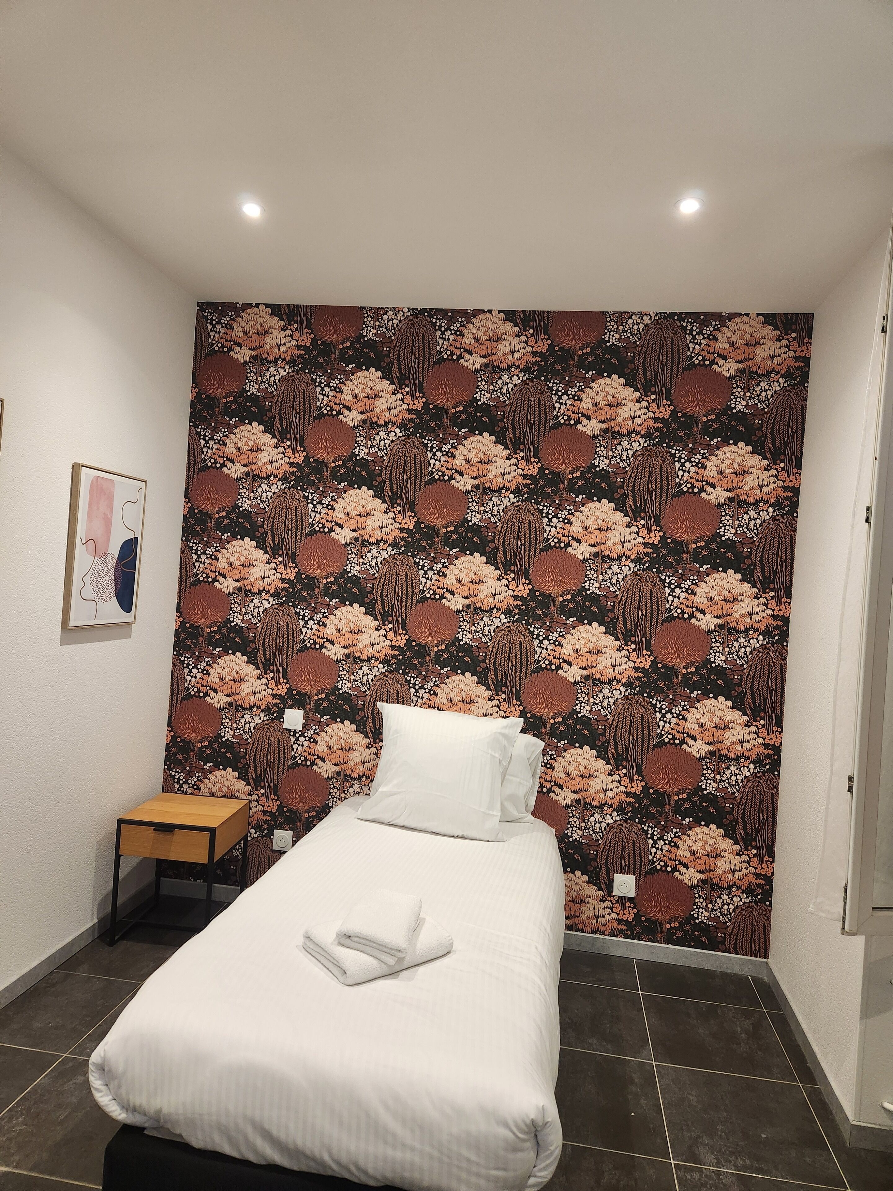 A bedroom with a bed, a small wooden nightstand, a wall-mounted light, and a patterned wallpaper.