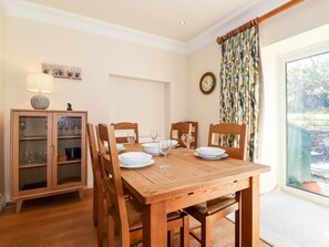 Un coin repas avec une table en bois dressée pour quatre personnes, entourée de chaises, un meuble avec des portes en verre et une fenêtre avec des rideaux.