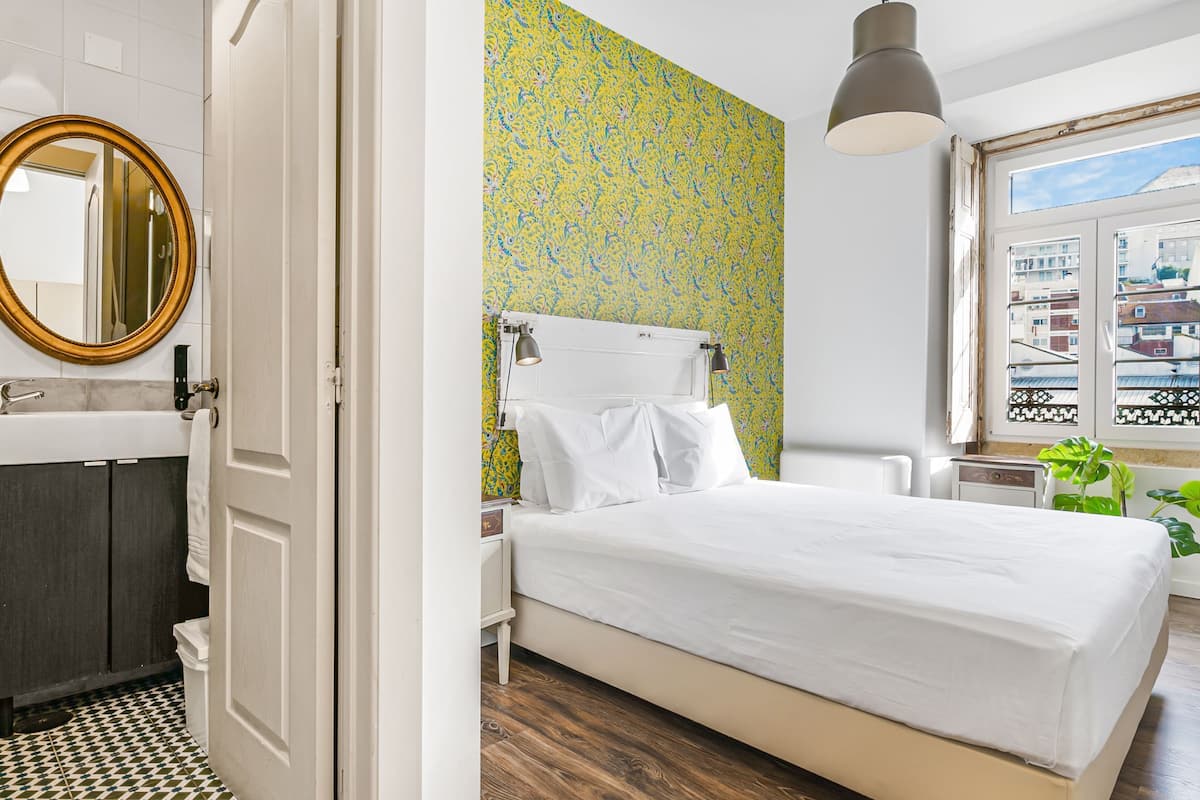 A bedroom with a bed, a nightstand, a mirror, a window with a view, and a bathroom with a sink and toilet.