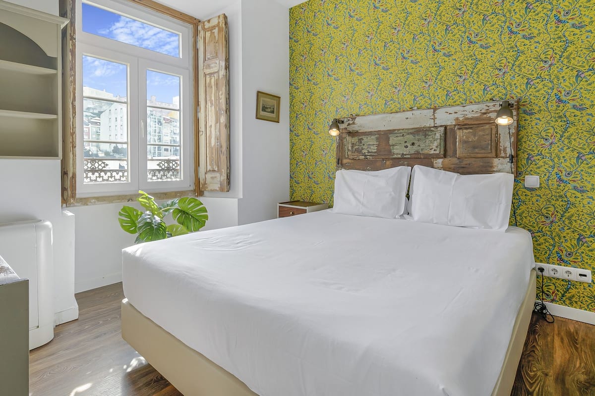 A bedroom with a large bed, a window with curtains, a plant, and a patterned wall.
