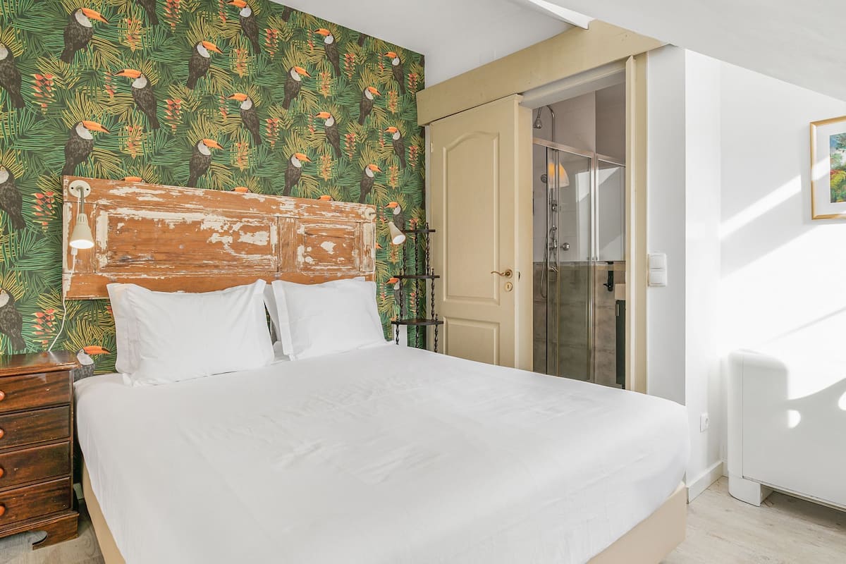 A bedroom with a bed, a wooden headboard, a nightstand, a dresser, a shower, and a door leading to a bathroom.