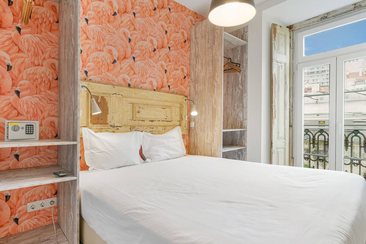 A bedroom with a wooden headboard, white bedding, a radio on a shelf, and a view of the city through a window.