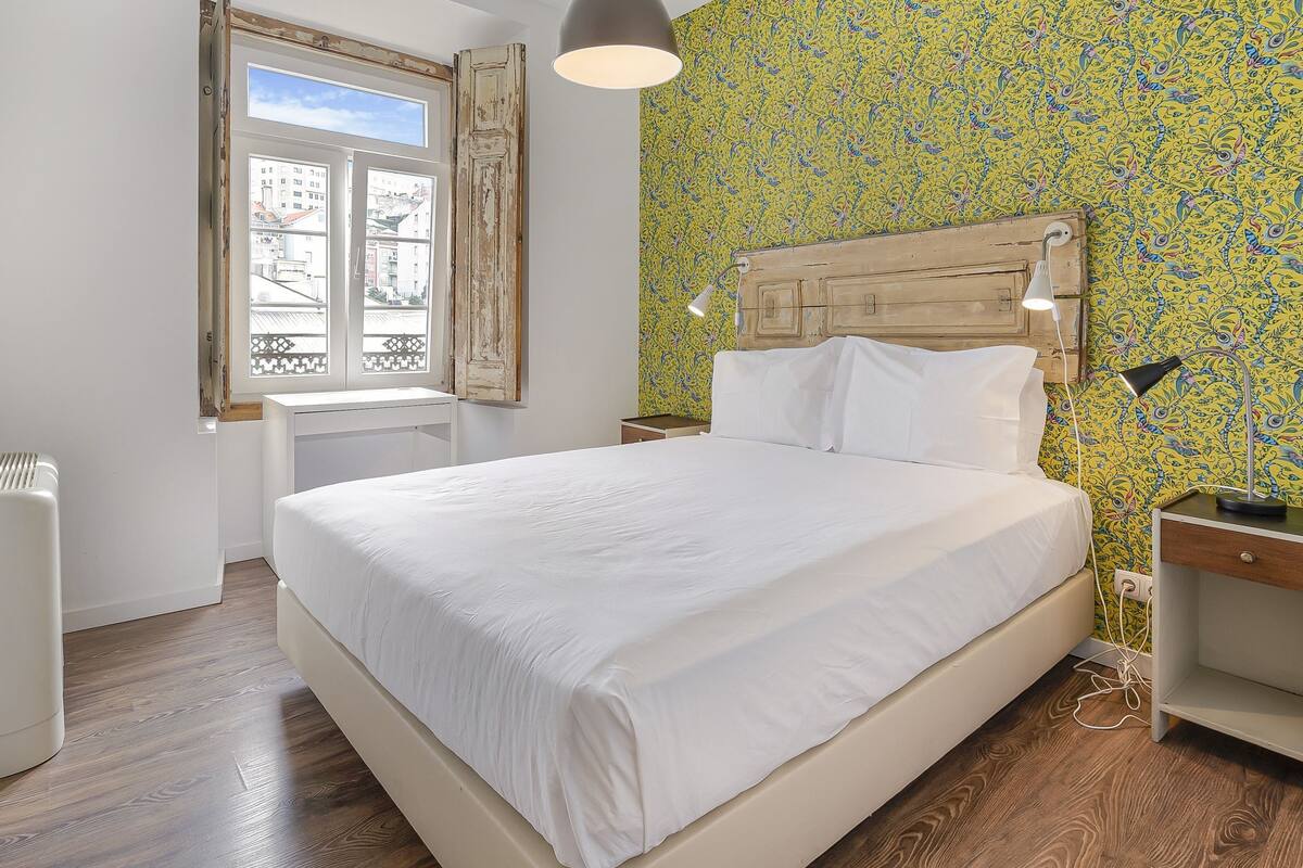 A bedroom with a large bed, a wooden headboard, a window with curtains, a bedside table with a lamp, and a wall with a floral pattern.