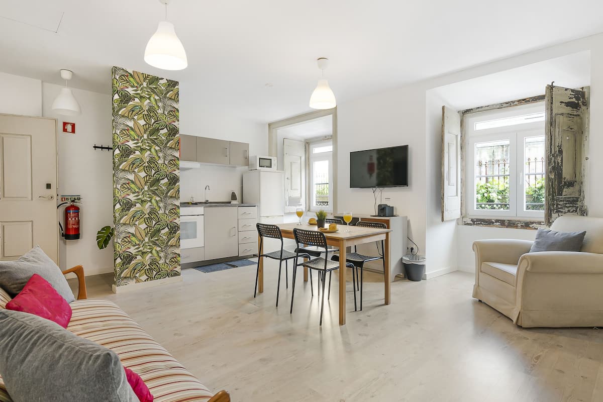 A modern living room with a sofa, a dining table with chairs, a kitchen area, and a television mounted on the wall.