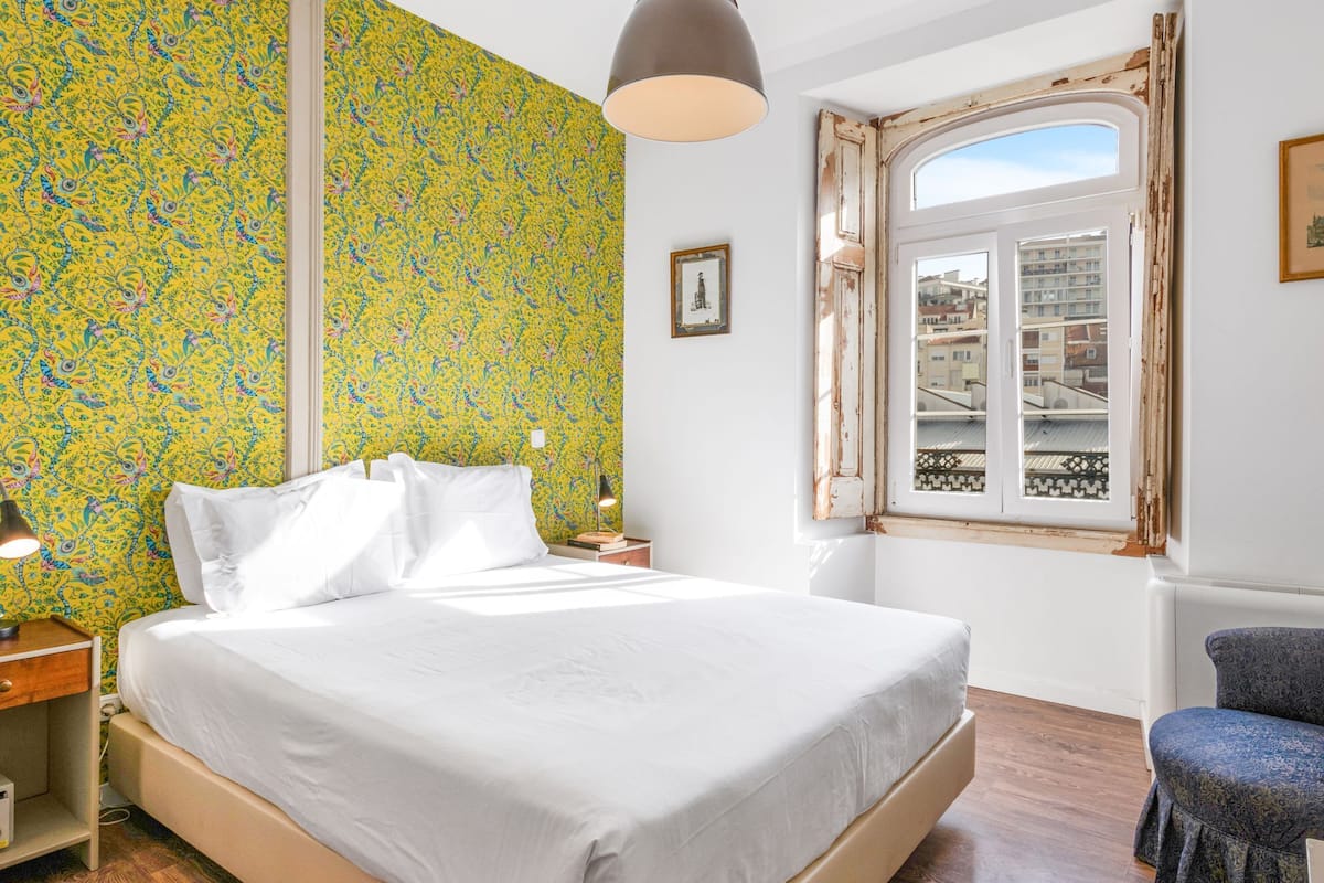 A bedroom with a large bed, a nightstand, a lamp, a window with a view of buildings, and a chair.