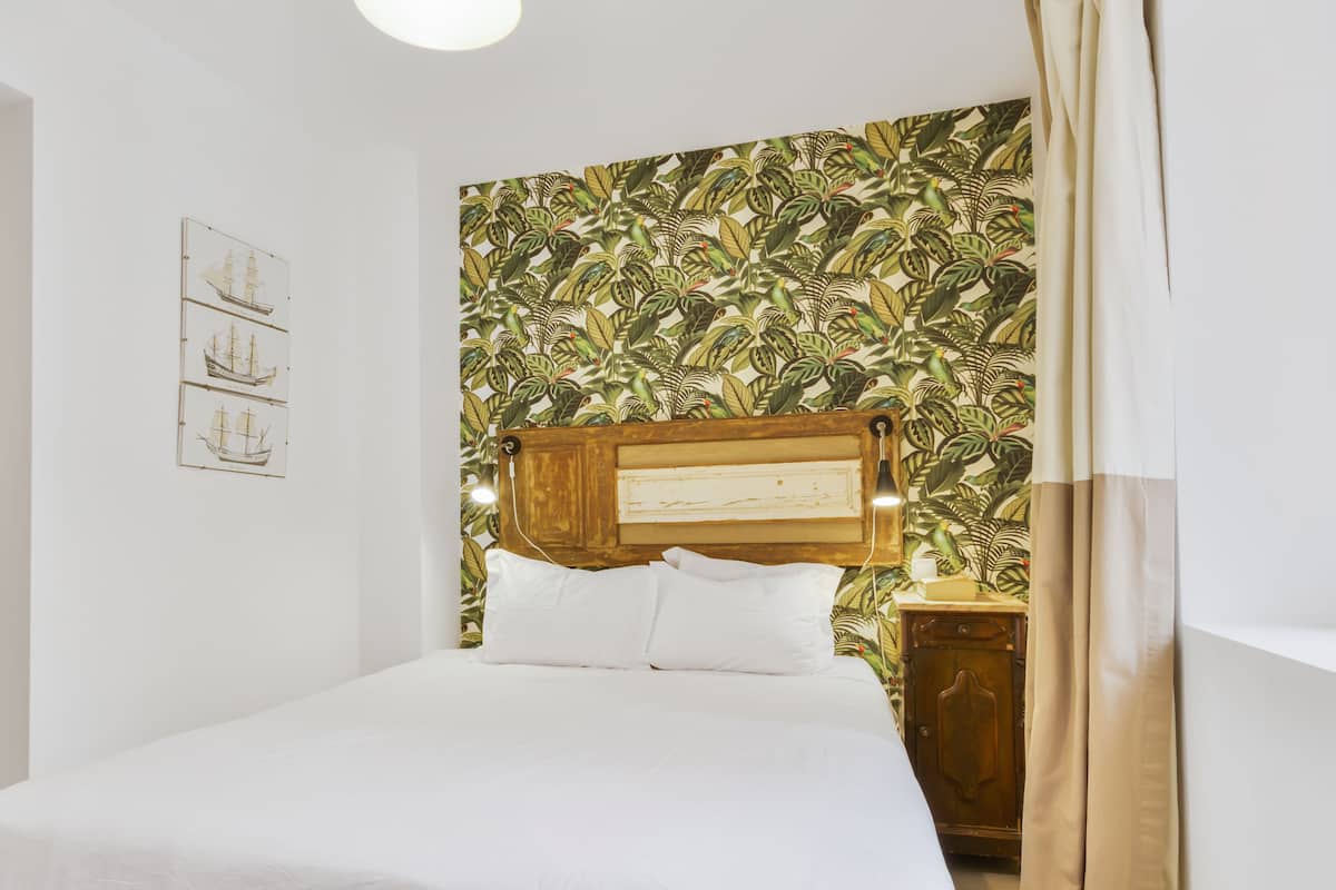 A neatly made bed with white linens, a wooden headboard with a plaque, and a tropical leaf-patterned wallpaper.