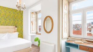 Una camera da letto con un letto, una cassettiera, uno specchio, un termosifone e una finestra con vista sugli edifici.