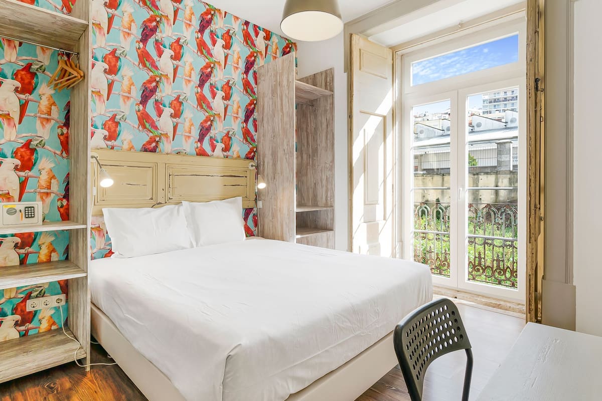 A bedroom with a bed, a wooden headboard, a wardrobe with a colorful bird wallpaper, and a window with a view of buildings.