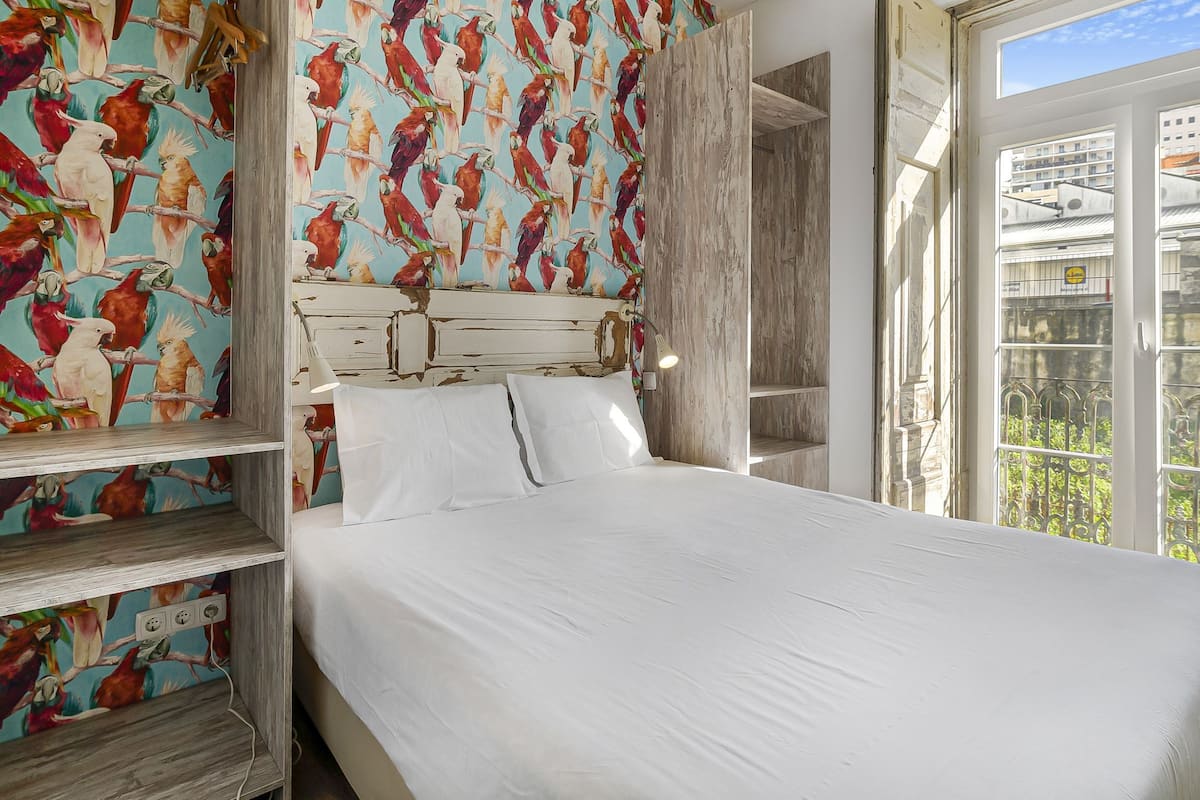 A bedroom with a bed, white bedding, a wooden headboard, a colorful parrot wallpaper, and a view of buildings through a window.